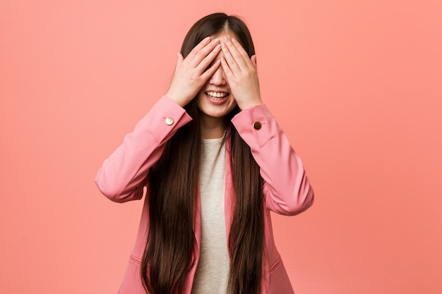 La mujer china joven del negocio que lleva el traje rosado cubre los ojos con las manos, sonríe ampliamente esperando una sorpresa.