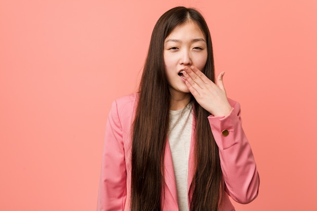 Mujer china joven del negocio que lleva el traje rosado que bosteza mostrando un gesto cansado que cubre la boca con su mano.