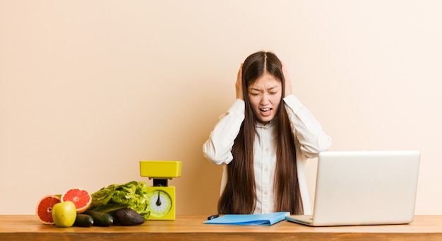 Mujer china joven nutricionista que trabaja con su computadora portátil coning oídos con las manos tratando de no escuchar un sonido demasiado fuerte.