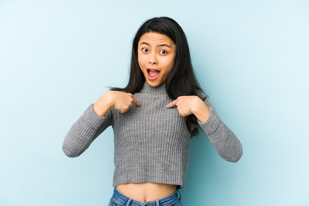 La mujer china joven en una pared azul sorprendió señalar con el dedo, sonriendo ampliamente.