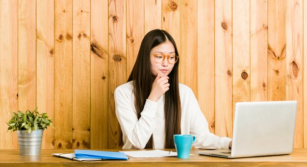 Mujer china joven que estudia en su escritorio que mira de lado con expresión dudosa y escéptica.