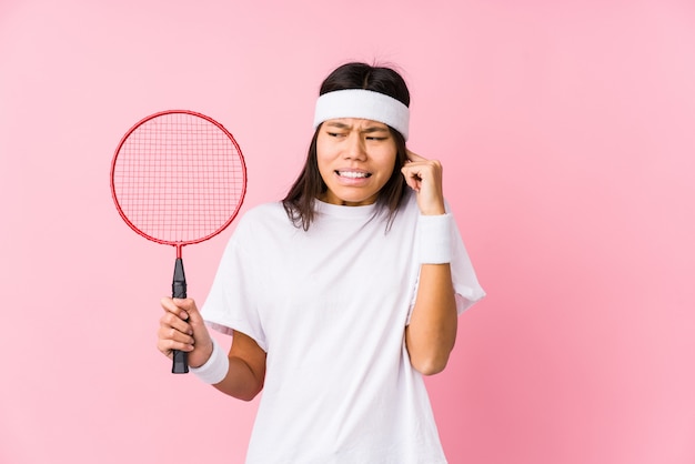 Mujer china joven que juega a bádminton en una pared rosada que cubre los oídos con las manos.