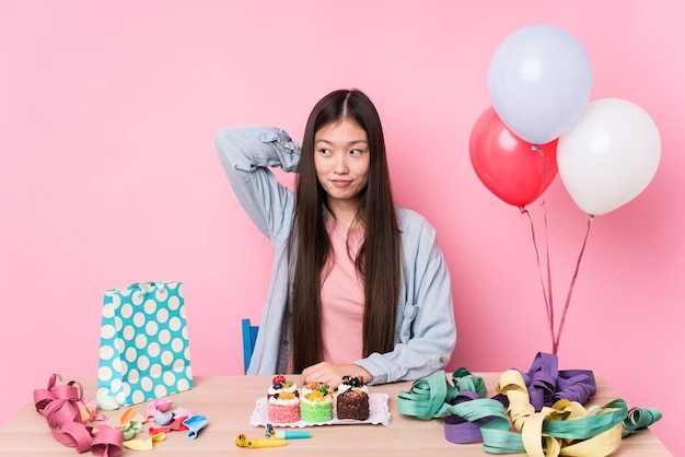 Mujer china joven que organiza un cumpleaños aislado tocando la parte posterior de la cabeza, pensando y haciendo una elección.