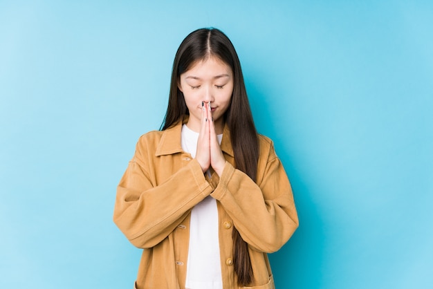 La mujer china joven que presenta en una pared azul aislada que lleva a cabo las manos en ruega cerca de boca, se siente confiada.