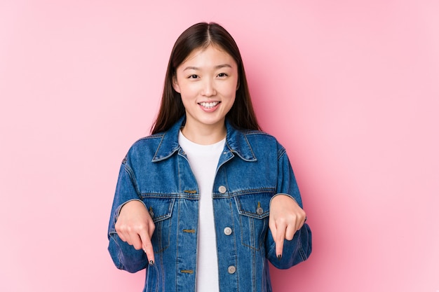 La mujer china joven que presenta en una pared rosada aisló puntos abajo con los dedos, sensación positiva.