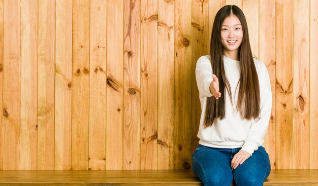 Mujer china joven que se sienta en un lugar de madera que estira la mano en la cámara en gesto del saludo.