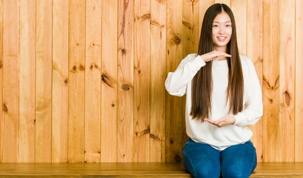 Mujer china joven que se sienta en un lugar de madera que lleva a cabo algo con ambas manos, presentación del producto.