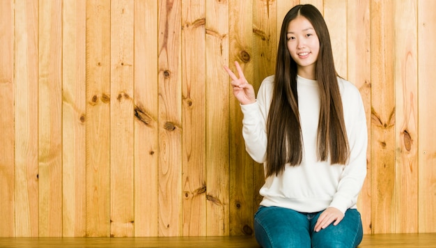 Mujer china joven que se sienta en un lugar de madera que muestra la muestra de la victoria y que sonríe ampliamente.