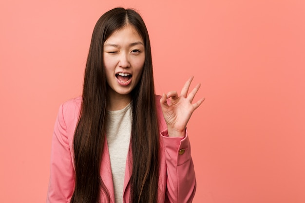 La mujer china del negocio joven que lleva el traje rosado guiña un ojo y sostiene un gesto aceptable con la mano.