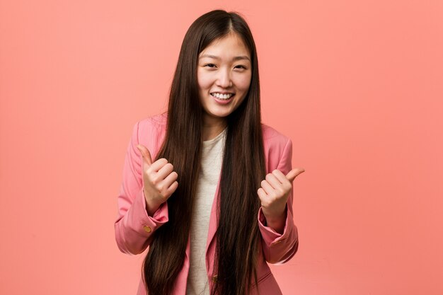 Mujer china del negocio joven que lleva el traje rosado que levanta ambos pulgares para arriba, sonriendo y confiado.