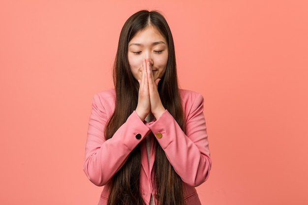 Mujer china de negocios joven con traje rosa cogidos de la mano en oración cerca de la boca, se siente segura.