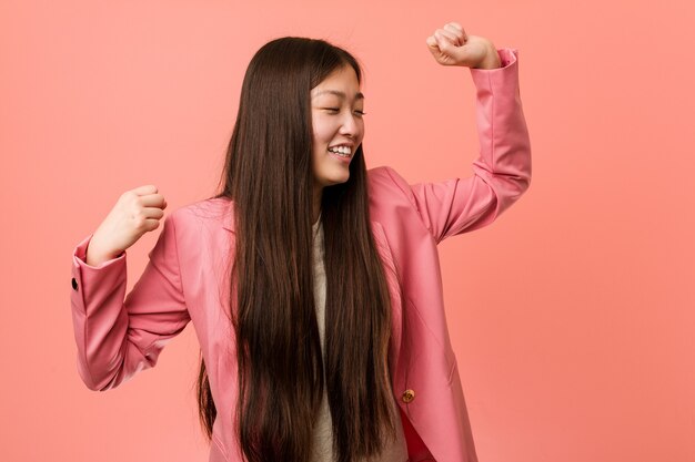 Mujer china de negocios joven vestida con traje rosa celebrando un día especial, salta y levanta los brazos con energía.