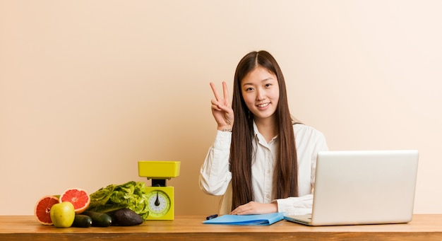 Mujer china del nutricionista joven que trabaja con su computadora portátil que muestra la muestra de la victoria y que sonríe ampliamente.