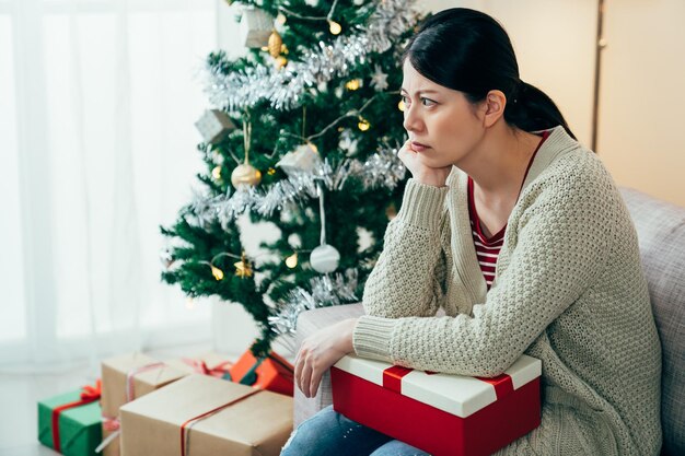 mujer china sentada en un silencio malhumorado con una caja no está contenta con el regalo de Navidad barato que recibió. una chica asiática está mirando hacia otro lado con ira porque su novio no pasó tiempo con ella en las vacaciones