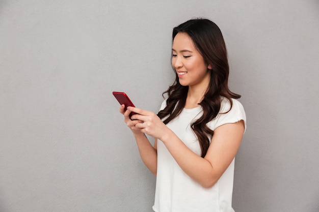 Mujer china sonriente contenta en camiseta casual escribiendo mensajes de texto o desplazando la red social con un teléfono inteligente, aislado sobre una pared gris