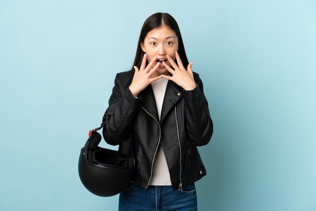 Mujer china sosteniendo un casco de moto sobre pared azul aislado con expresión facial sorpresa