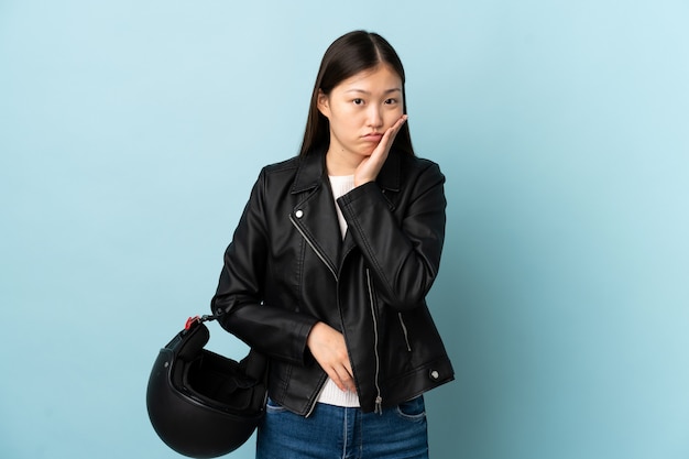 Mujer China sosteniendo un casco de motocicleta sobre una pared azul aislada infeliz y frustrada