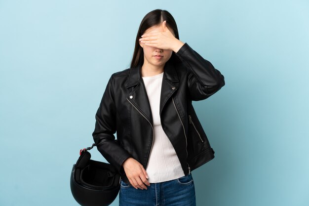 Mujer china sosteniendo un casco de motocicleta sobre la pared azul aislada que cubre los ojos con las manos