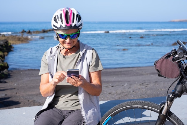Mujer ciclista senior sonriente con casco sentado en la playa con bicicleta eléctrica usando un teléfono inteligente
