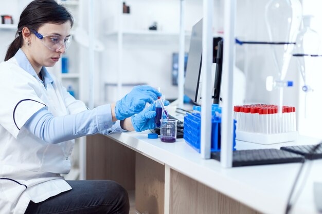 Mujer científica en la industria de la salud tomando muestra de tubo de ensayo con pipeta de vidrio. Científico químico en laboratorio estéril utilizando tecnología moderna para probar líquido de microbiología.