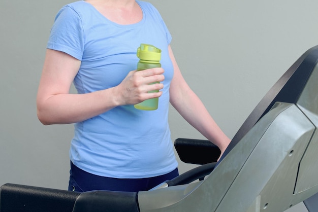 La mujer se para en una cinta de correr en el gimnasio y sostiene una botella deportiva con agua