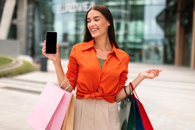 Mujer cliente alegre mostrando pantalla en blanco de teléfono celular en un área urbana