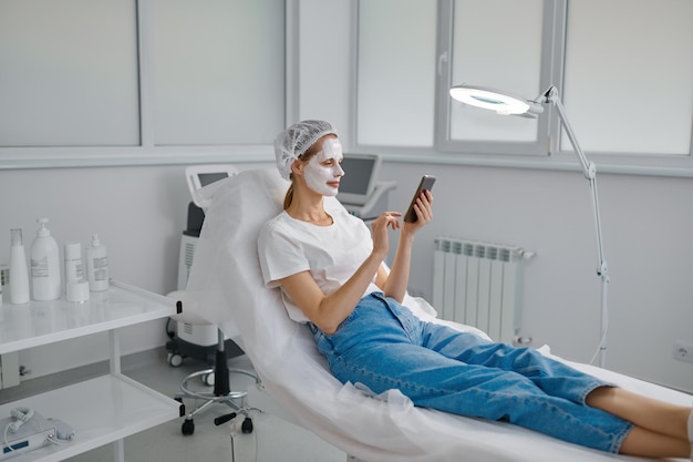 Mujer cliente con mascarilla de arcilla descansando y usando teléfono móvil para chatear en redes sociales. Servicio de cosmología y concepto de tratamiento de belleza.