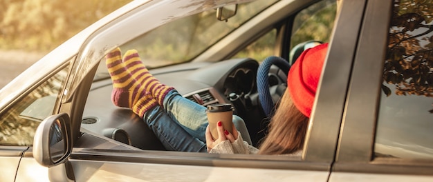 Mujer en un coche en cálidos calcetines de lana amarillo está sosteniendo una taza de café en las manos al atardecer