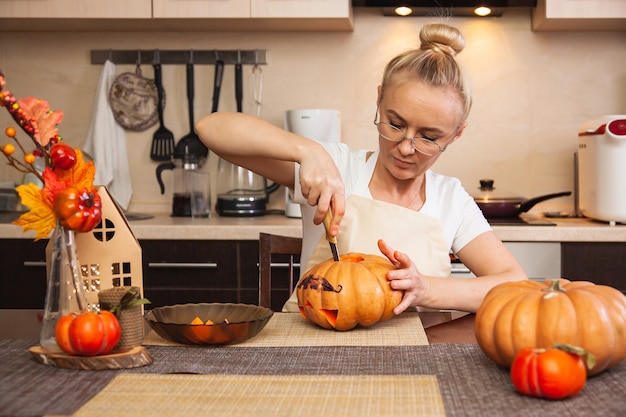 Mujer en la cocina talla una calabaza para Halloween en una habitación con decoración otoñal y una casa de lámparas. Acogedora casa y preparándose para Halloween.