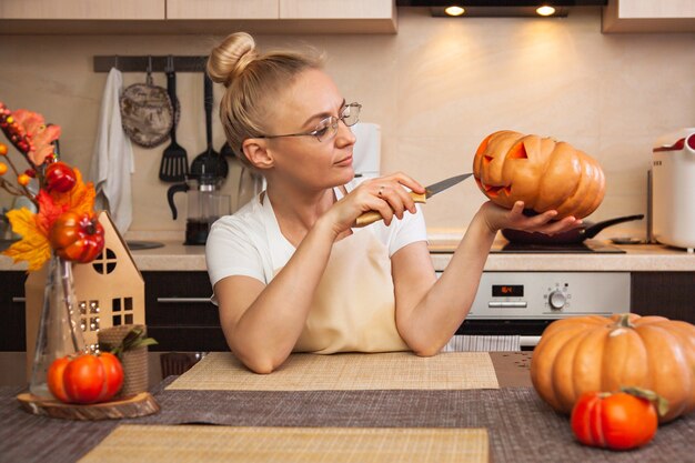 Mujer en la cocina talla una calabaza para Halloween en una habitación con decoración otoñal y una casa de lámparas. Acogedora casa y preparándose para Halloween.