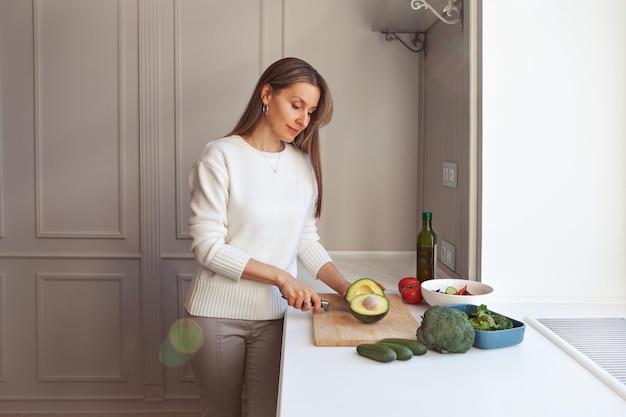 Mujer cocinando ensalada vegetariana con verduras frescas. Modelo de corte con cuchillo aguacates, brócoli, ensalada de col rizada, pepinos y tomates en cocina blanca