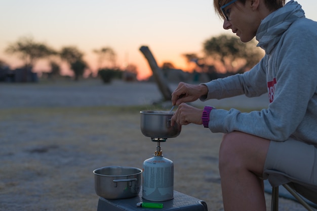 Mujer cocinando con estufa de gas en camping al atardecer. quemador de gas, olla y humo del agua hirviendo.