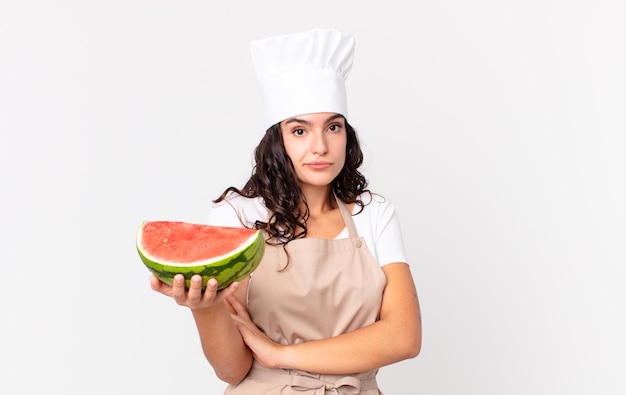 Mujer cocinera bonita hispana encogiéndose de hombros sintiéndose confundida e insegura y sosteniendo una sandía