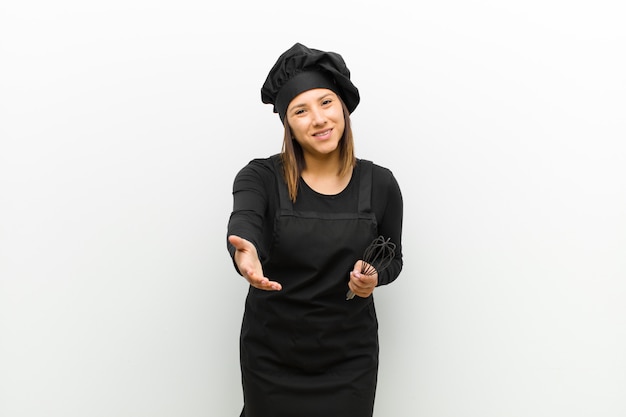 Mujer cocinera sonriente, feliz, confiada y amigable, ofreciendo un apretón de manos para cerrar un trato, cooperando contra la pared blanca