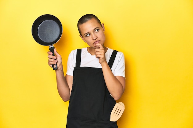 Mujer cocinera sosteniendo una sartén mirando hacia los lados con expresión dudosa y escéptica