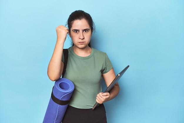 Mujer con colchoneta y computadora portátil en azul mostrando el puño a la cámara expresión facial agresiva