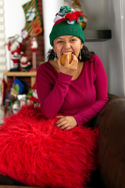 Mujer colombiana en Navidad con sombrero de Navidad sentado en el sofá comiendo donut comida típica colombiana