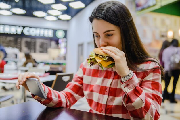 Foto mujer come humburger mientras busca el teléfono