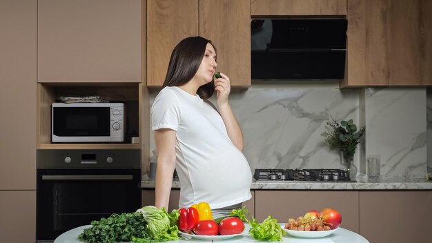 Mujer come verduras acariciando el vientre embarazado en la cocina