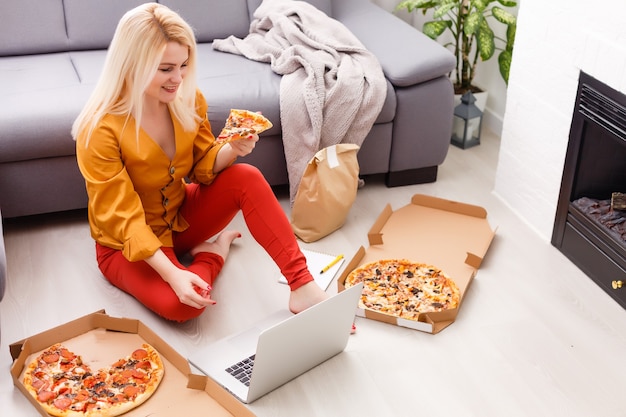 Mujer de comercio electrónico. Mujer joven con computadora para compras en línea, sentada en el suelo y comiendo pizza.