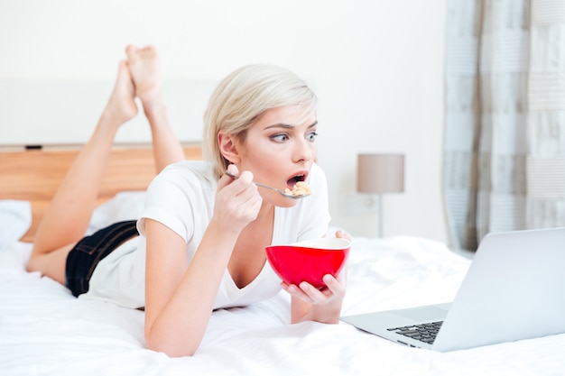 Mujer comiendo cereal en la cama y viendo una película en la computadora portátil en casa