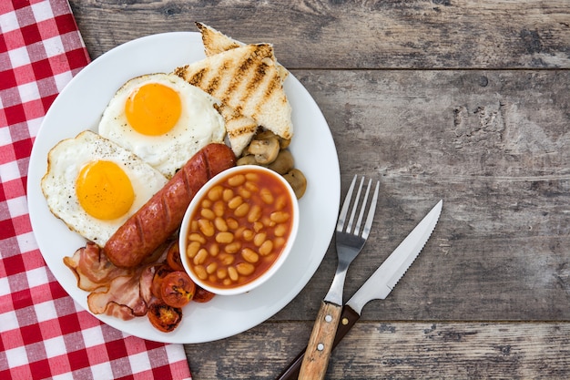 Mujer comiendo un desayuno tradicional inglés con huevos fritos, salchichas, frijoles, champiñones, tomates a la parrilla, tocino en la mesa de madera