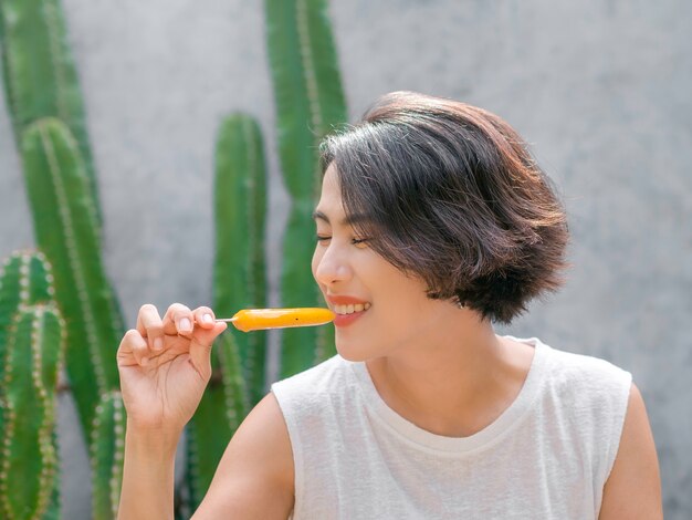 Foto mujer comiendo paletas heladas. feliz hermosa mujer asiática con camisa sin mangas blanca casual con paleta amarilla, al aire libre. mujer sonriente disfrutando de polo de hielo en verano.