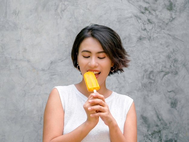 Mujer comiendo paletas heladas. Feliz hermosa mujer asiática de pelo corto con camisa blanca sin mangas casual con paleta amarilla sobre fondo gris muro de hormigón en verano.