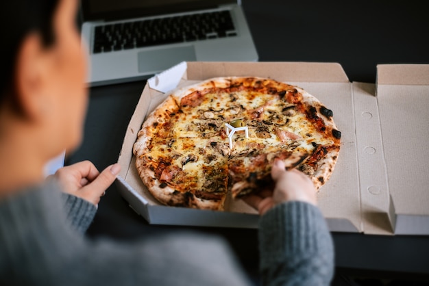 Mujer comiendo pizza mientras trabaja en la computadora portátil.