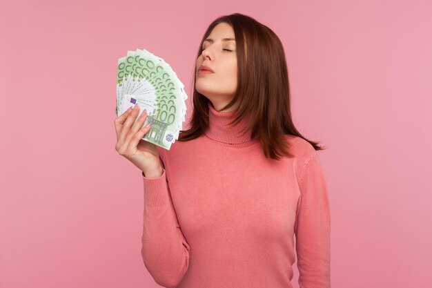 Mujer complacida satisfecha con cabello castaño en suéter rosa que huele a abanico de cientos de euros en la mano, disfrutando del dinero, la riqueza. Disparo de estudio interior aislado sobre fondo rosa