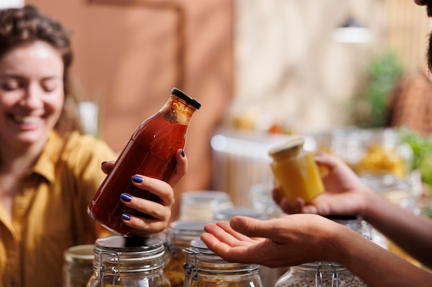 Una mujer compra en una tienda de comestibles sin desperdicio, mira productos a granel en envases reutilizables mientras es asistida por un feliz tendero. Comerciante en una tienda ecológica recomendando alimentos al cliente, de cerca