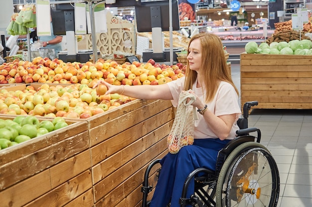 Mujer compradora que usa silla de ruedas elige manzanas en un supermercado y las pone en una bolsa de malla