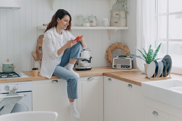 Mujer comprando comestibles en línea mediante una aplicación de teléfono móvil sentada en una cocina moderna en casa Comercio electrónico