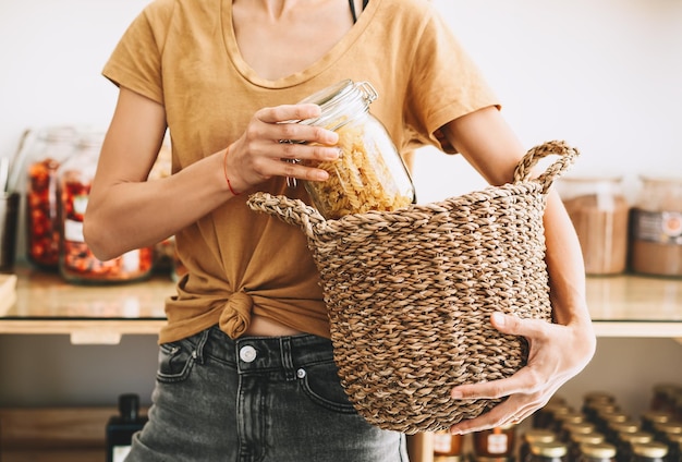 Mujer comprando productos en una tienda de comestibles sin plástico Frasco de vidrio con pasta en manos de una persona en una tienda de desperdicio cero Niña con canasta de mimbre hace compras conscientes Pequeñas empresas sostenibles